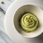 Pistachio cream with a smooth swirl, topped with crushed pistachios and seeds, served in a speckled ceramic bowl on a marble board.