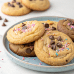 Plate of Crumbl cookies with chocolate chips and pink, white, and blue sprinkles, with additional cookies and chocolate chips in the background.
