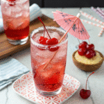 Sparkling Shirley Temple drink with ice, maraschino cherries, a sugared rim, and a pink cocktail umbrella, served on a patterned plate.
