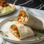 Two buffalo chicken wrap halves on a white plate with a gold rim, showing spicy chicken, shredded lettuce, cheese, and ranch dressing against a marble countertop.